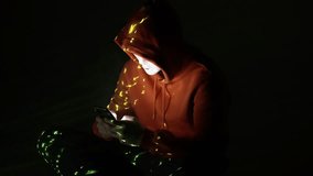 In a pitch-black room, a hooded figure in a white mask stares at a smartphone, illuminated by scattered yellow digital fragments, evoking cyber secrecy and mobile-based intrusion attempts - Powered by Shutterstock - Get 15% off with code: PIKWIZARD15