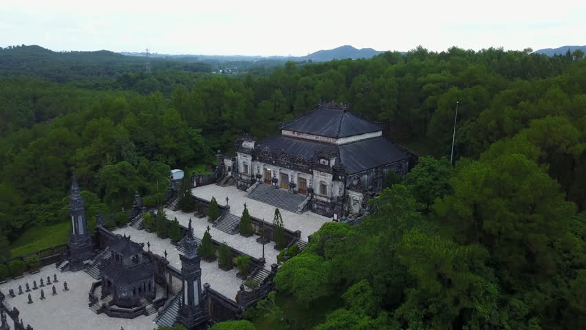 An expansive view pulling back from the Mausoleum of Emperor Khai Dinh, situated amidst the lush landscape of Hue, Vietnam, showcasing its unique architecture and surroundings.