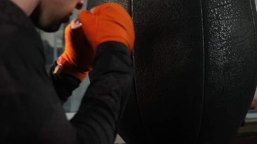 Boxer works with punching bag practicing punches in hall. Guy in bandages hones skills and strengthens muscles concentrating on constuction surface