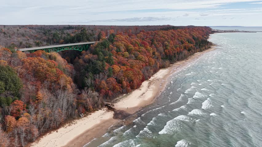 Aerial view of a secluded Lake Michigan shoreline lined with colorful autumn trees and a scenic coastal highway bridge in the Upper Peninsula