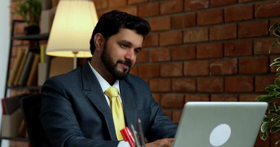 Happy Indian Asian handsome young businessman or CEO in formal corporate attire with beard, working on laptop computer in cozy modern office with terracotta brick wall