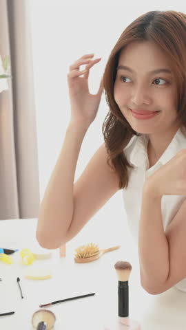 Young Asian woman slowly touching lips with lipstick in graceful motion, showing elegance and concentration while enjoying beauty moment on makeup table in bright room, Vertical