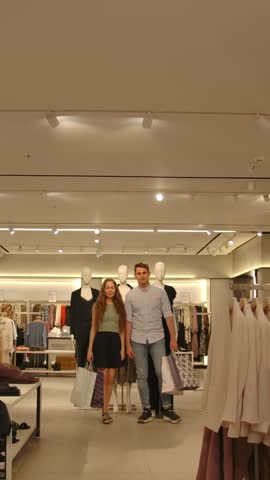 Happy married couple with shopping packages approaches the camera with a smile. Cute loving family in a shirt bought in a new modern clothing store. Middle shot. Vertical shot.