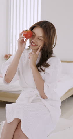 Young Asian woman smiling while holding fresh tomato in spa robe, promoting natural beauty and healthy skin through fruits in casual beauty routine at home, Vertical