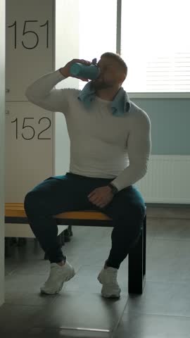 A man drinks water and towels off in the locker room after training. Rest from power loads. Window and lockers in the background. Slow motion panning shot. Vertical shot.