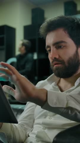 Young caucasian man checking social media on a tablet PC, while waiting for his turn to cut in a barbershop. Barber girl making hairstyle to a client with pink hair at the background. Vertical shot.