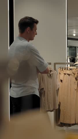 Caucasian woman in the store asks the guy's advice choosing between two dresses. The couple in love looking for clothes in store of modern clothing for shopping. Slow motion wide shot. Vertical shot.
