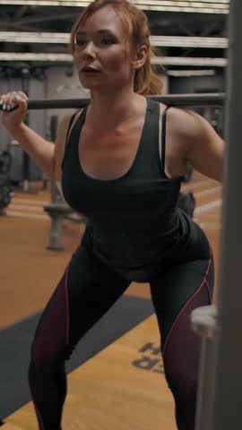 Sexy athlete woman doing barbell squats in slow motion. interior of a modern sports hall with dumbbells and gym equipment in the background. Vertical shot.