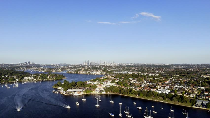 A breathtaking panoramic view of a Sydney waterfront, showcasing numerous boats on the water, lush green peninsulas dotted with houses, and the iconic city skyline