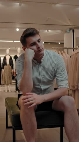 A bored man sits on a bench in a clothing store. He is tired of waiting for his girlfriend to choose a fashionable outfit. Vertical shot.