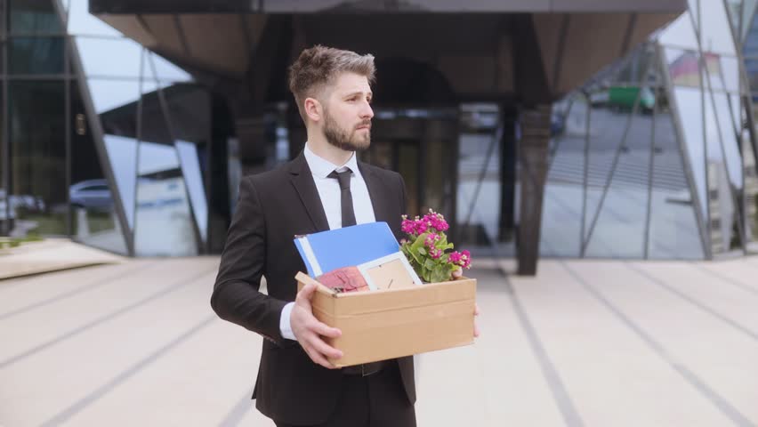 A white collar worker stands outside a glass office building holding a box of personal items and flowers. He shows signs of uncertainty after receiving layoff news.