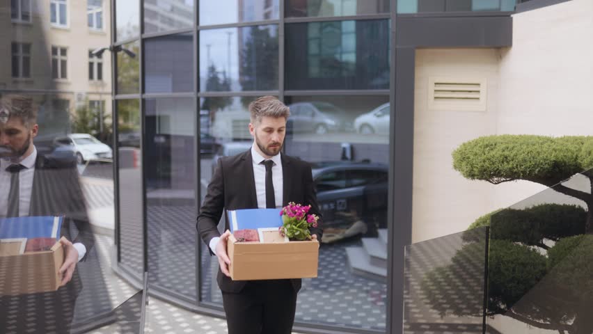 Upset young man in business suit shouts in shock outside corporate building after losing job. Cardboard box in hand symbolizes sudden layoff and emotional stress.