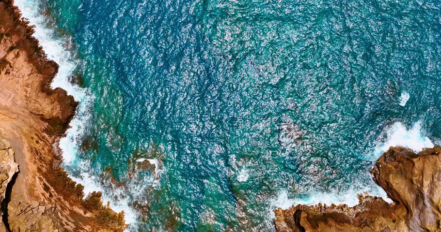 Aerial top-down view of the calm blue waters of the Pacific Ocean off the rocky lava shores of Hawaii.