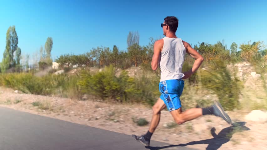 A young athlete in a tank top and shorts races down a countryside asphalt road at high speed. The road leads to the sea, with bushes and scattered trees lining the edges under a bright blue sky.