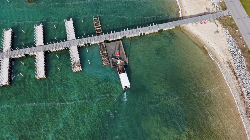 Aerial drone view of a wooden pier extending into emerald-green waters along a sandy Lake Michigan shoreline in the Upper Peninsula