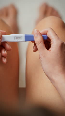 A young girl sitting in the bathroom checks pregnancy test. She looks at the test where the indicator shows two stripes, which means she is pregnant. The actions are accompanied by emotional reaction
