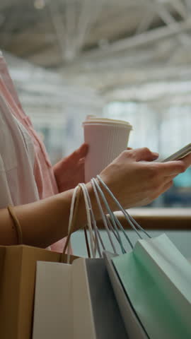 A business woman is holding a coffee cup and a phone while stopping to browse in a department store. Shopping bags hang from her wrist, indicating a day of exploration and retail therapy