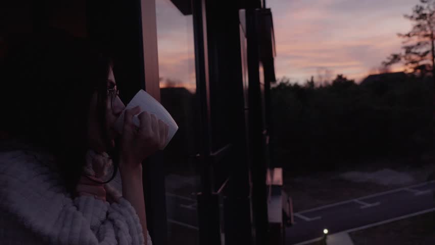 Young woman wrapped in a blanket enjoying a cup of coffee or tea on her balcony at sunset, slow motion