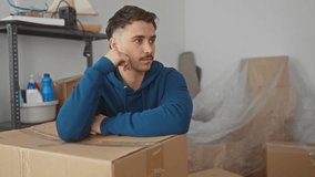 Young man in blue hoodie leaning on cardboard box in new apartment surrounded by unpacked items, symbolizing transition and fresh beginnings in a modern residential space. - Powered by Shutterstock - Get 15% off with code: PIKWIZARD15