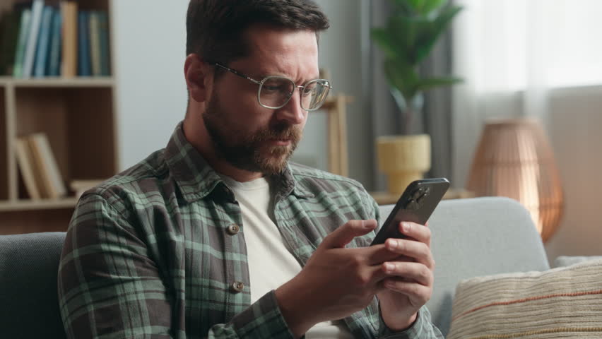 Caucasian adult man male guy at home living room nervous looking at mobile phone bad news amazed shocked close mouth with hand message from bank stressed smartphone notification online error scam
