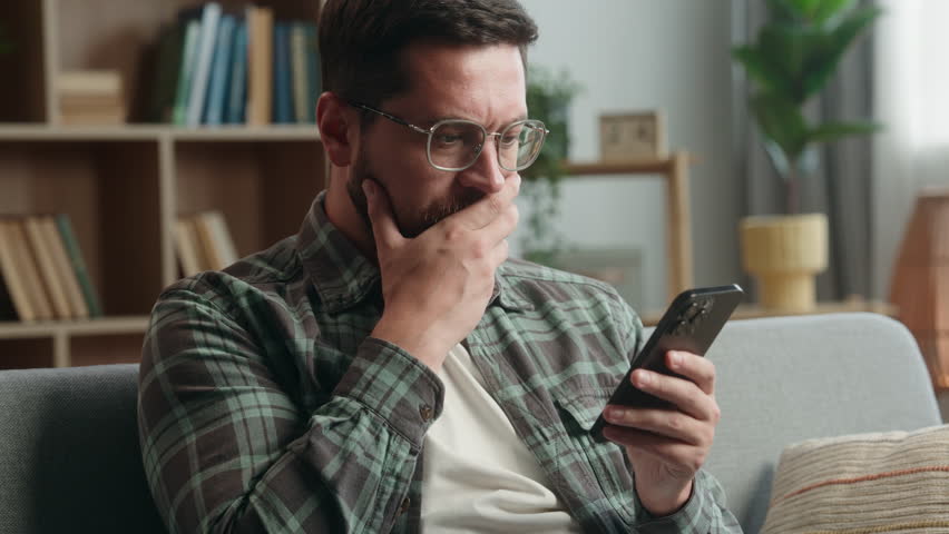 Caucasian adult man male guy at home living room nervous looking at mobile phone bad news amazed shocked close mouth with hand message from bank stressed smartphone notification online error scam