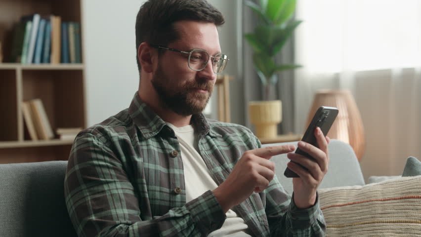 Smiling happy relaxed middle-aged man at home living room holding cell mobile phone using smartphone on couch online scrolling social media male guy checking apps buying shopping texting messages