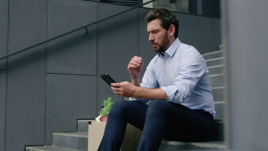 Serious fired unemployed Caucasian middle-aged businessman using smartphone searching work vacancy online thinking business man male guy in city with mobile phone with dismissal box dismiss lost job