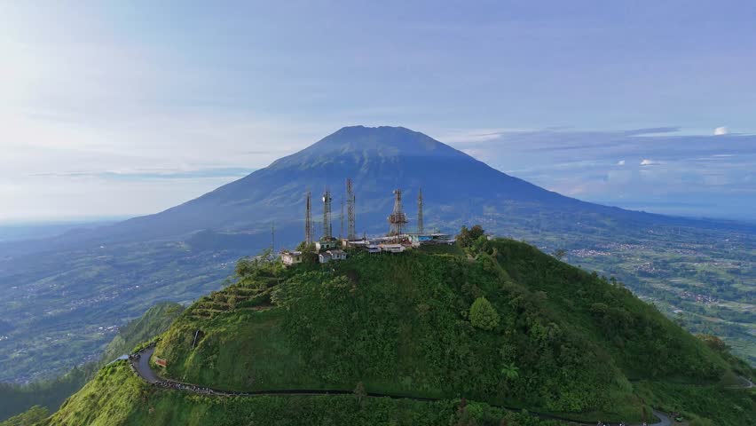 Drone flying close to mountain peak with buildings on it. Mount Telomoyo with Mount Merbabu in the background. Indonesia.
