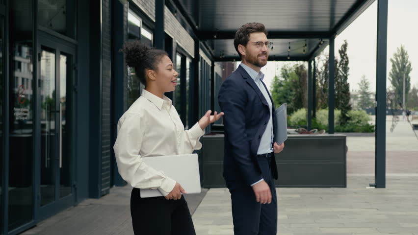 Two multiracial business people colleagues leaving office together walking outside in city company building leave African American businesswoman woman female with Caucasian man businessman guy talking
