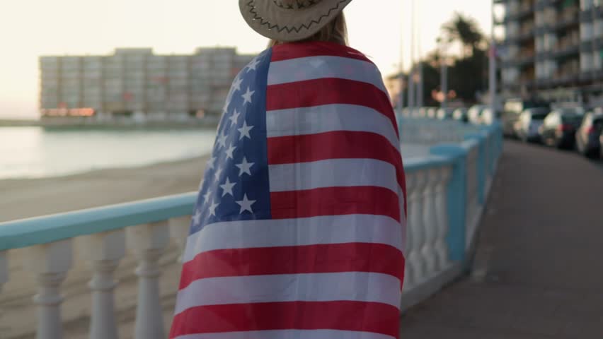 Patriotic blonde woman walking boardwalk at sunset, wrapped in american flag symbolizing summer freedom and national spirit.Usa celebrate 4th of july. Independence day concept