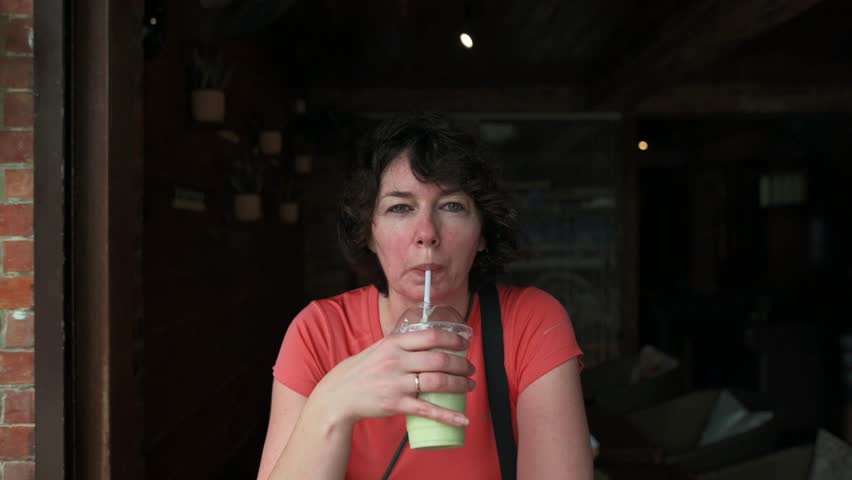 Woman in orange shirt holding plastic cup drinking green smoothie with straw while standing at entrance dimly lit rustic cafe with brick wall hanging potted plants background drinking green smoothie 