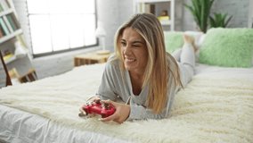 Woman playing video games in cozy bedroom, lying on bed with red game controller, featuring blonde hair, casual attire, smiling expression, modern home interior, and green pillows. - Powered by Shutterstock - Get 15% off with code: PIKWIZARD15