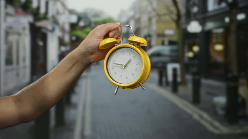 Male hand holding a yellow clock while pointing at it on a city street, suggesting urgency or time awareness against an urban backdrop.