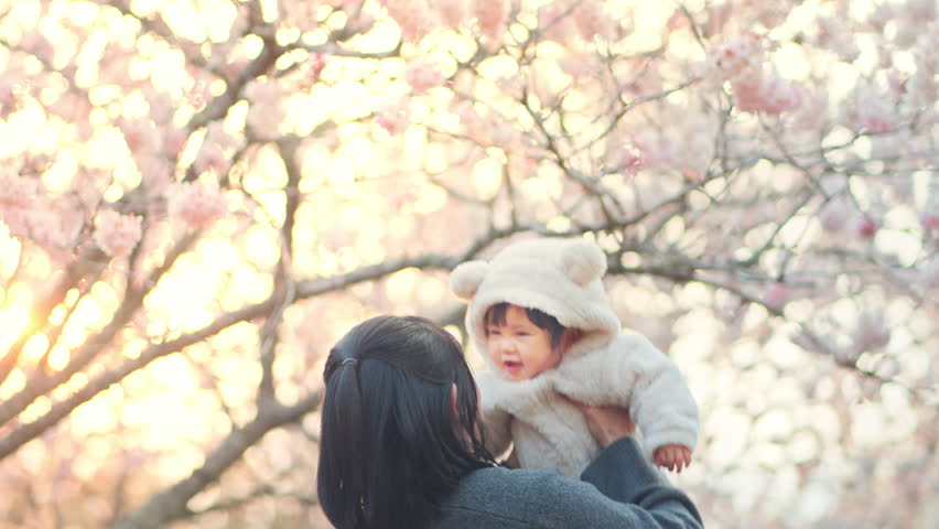 Happy little baby girl in her mother hands laughs and smiles over cherry blossom trees, Family playing together outside, Newborn spring concept