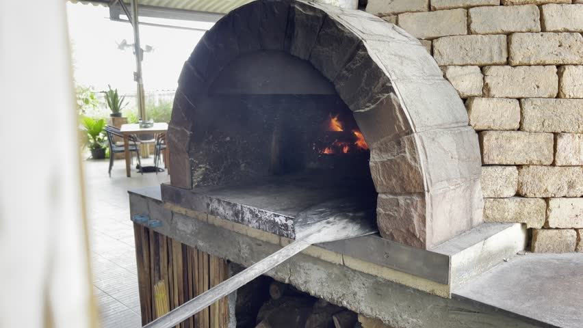 Close-up: chef placing pizza into traditional brick oven with open flame using long-handled pizza peel, authentic wood-fired cooking method, rustic preparation, hot fire, handmade pizza baking process