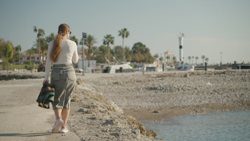 A woman walks along a seaside promenade toward a marina, holding a plaid jacket, with palm trees and boats in the background under bright sunlight