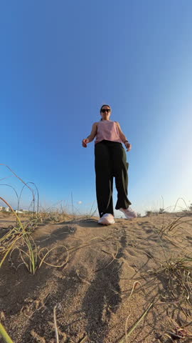 Vertical video. A worm's-eye view of a woman walking on sandy ground with sparse dry vegetation, her foot stepping forward, under a bright blue sky and warm sunlight