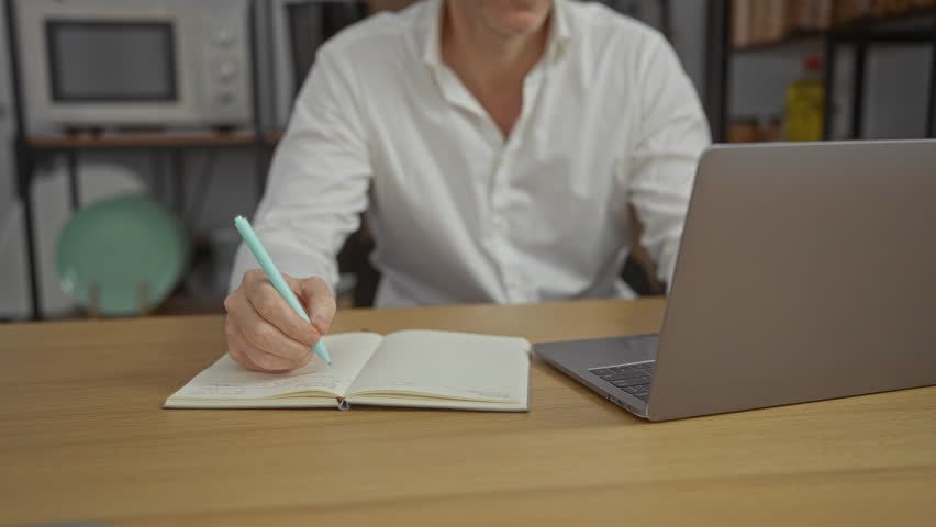 Middle-aged man writing notes in office setting with laptop on wooden desk and shelves in background. - Powered by Shutterstock - Get 15% off with code: PIKWIZARD15