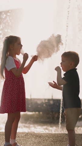 Little kids put spun sugar pieces into mouths refreshing near fountain in park slow motion. Tasty street dessert in amusement park. Sweet snack