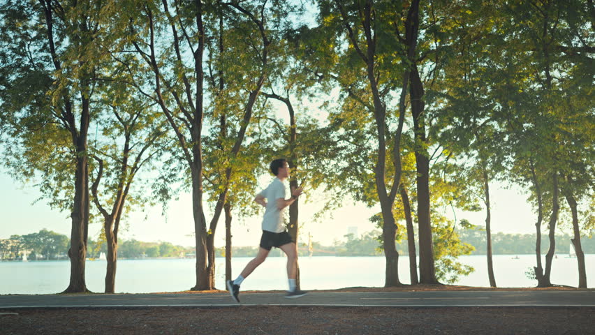 Side view - Attractive asian man jogging in the park on a background of trees with sunlight in sunset, Fitness Man Running, Training Run Workout.