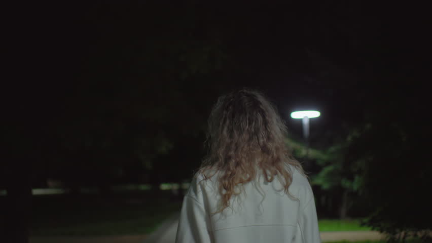 Back view young girl in white coat walking alone quietly at night along calm park walkway softly illuminated by gentle lights, creating atmosphere of peaceful solitude and quiet reflection