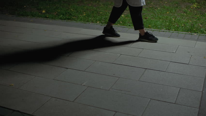 Leg view person walking along paved surface near grass, dim environment casting distinct shadows across walkway, capturing subtle motion in quiet night setting, footwear and gentle light