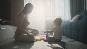 A parent sits across from a young child, both engaged in play with bright, colorful stacking toys. Sunlight streams through large windows, creating a warm atmosphere. - Powered by Shutterstock - Get 15% off with code: PIKWIZARD15