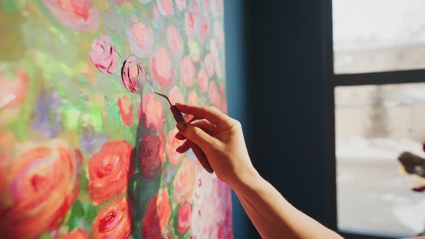 Talented artist carefully adds details to a vibrant painting of red roses, using a palette knife to apply thick strokes of paint on a large canvas in a naturally lit studio