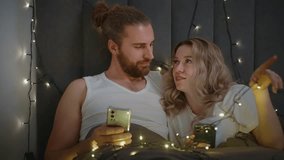 A couple is enjoying an intimate conversation in bed surrounded by string lights. They are both holding smartphones and sharing a warm moment together in a cozy atmosphere. - Powered by Shutterstock - Get 15% off with code: PIKWIZARD15