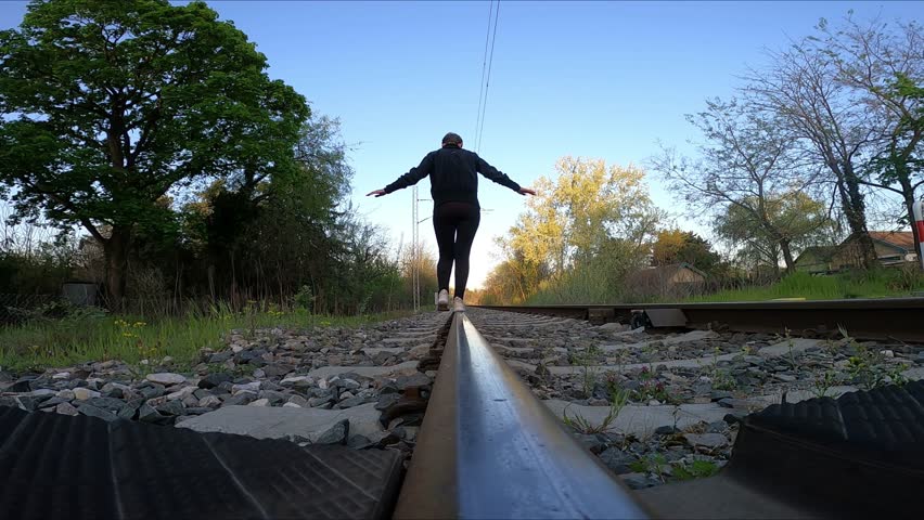 A woman in a black jumpsuit walks along the rails at dawn, balancing with her hands