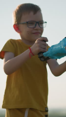 Young boy wearing a yellow shirt and shorts, with glasses, stands outdoors, holding a plastic container that releases bubbles into the air. in a bright, open field