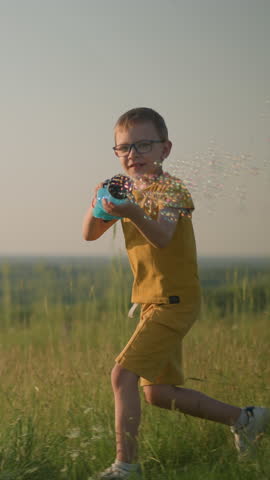 Young boy wearing a yellow shirt and glasses, smiling while holding a plastic bubble gun that releases bubbles. He stands in a grassy field under clear skies