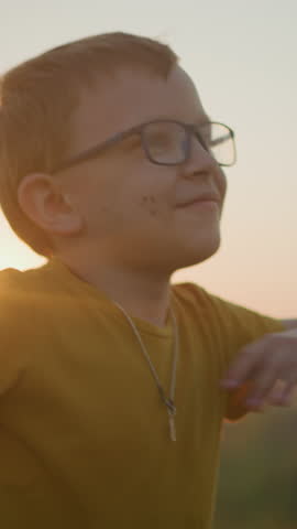 A young boy wearing a yellow shirt smiles shyly while moving his hands in a playful manner, standing in a sunlit grassy field during a serene sunset, capturing the essence of a joyful, carefree moment