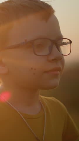 Close-up of a young boy wearing glasses and a yellow shirt, smiling shyly while playfully moving his hands in the soft, warm glow of the sunset. His gentle expression is a moment of joyful innocence
